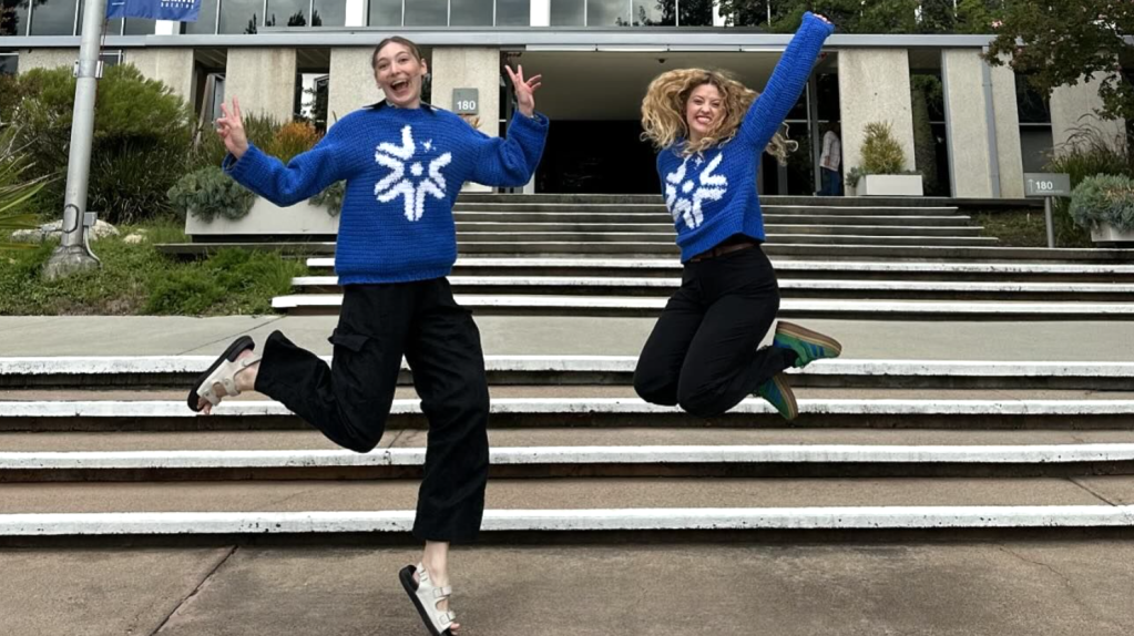 Two people wearing blue Europa Clipper sweaters jumping up with their arms raised.