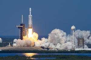 Europa Clipper lifts off from the launch pad atop a white rocket with two smaller side boosters. A bright, white and orange pillar of fire shoots from the bottom of the rocket stack. White smoke billows around the launch pad. The SpaceX fixed service structure is to the left of the rocket. A white water tower with the SpaceX logo is to the right. Farther right is a small, round, gray structure.