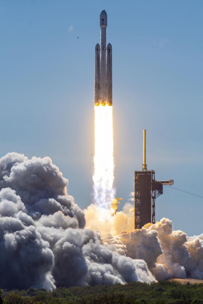 Europa Clipper lifts off from the launch pad atop a white rocket with two smaller side boosters. A bright, white and orange pillar of fire shoots from the bottom of the rocket stack. White smoke billows around the launch pad. The SpaceX fixed service structure is to the right of the rocket.