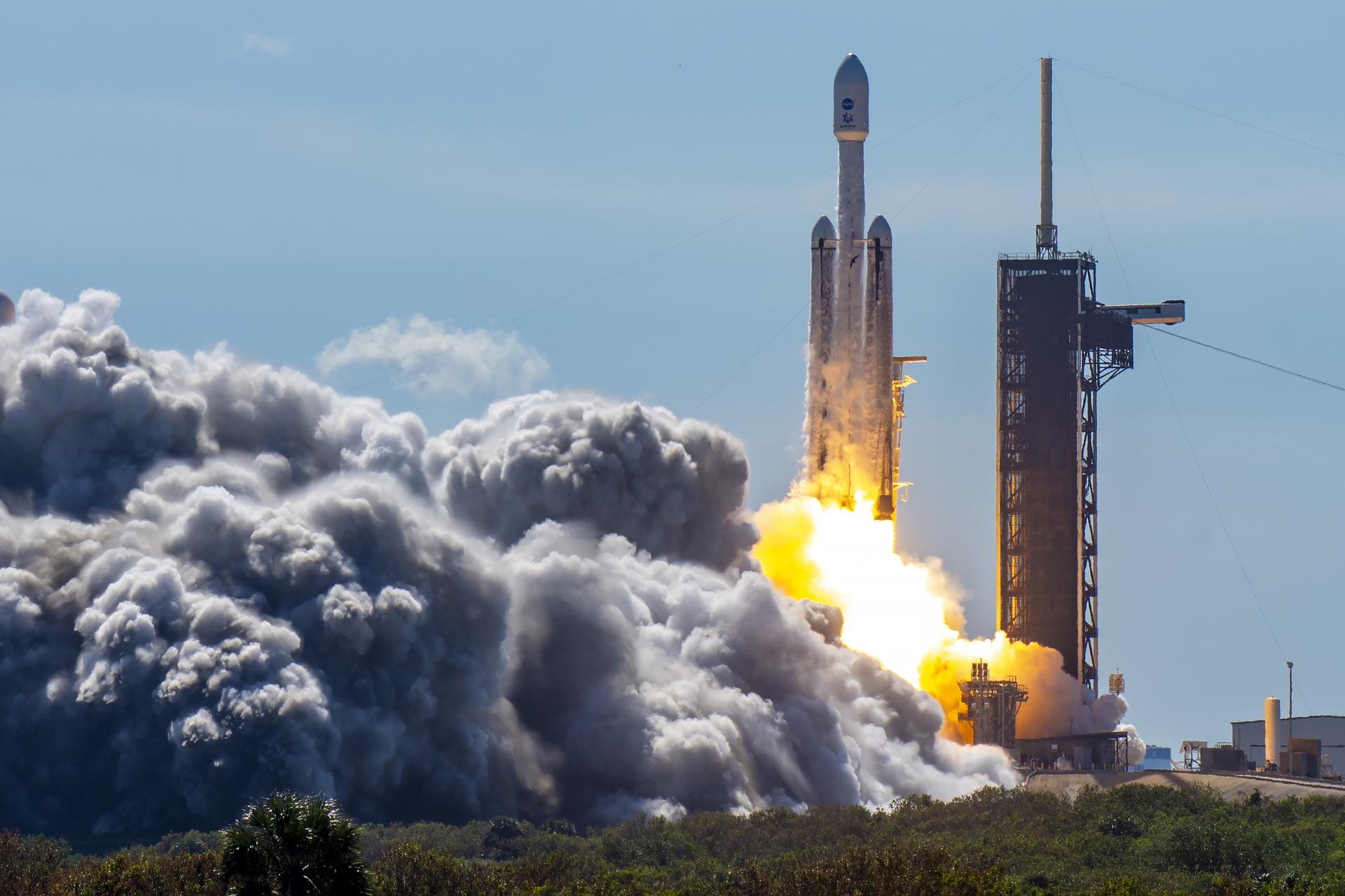 Europa Clipper lifts off from the launch pad atop a white rocket with two smaller side boosters. A bright, white and orange pillar of fire shoots from the bottom of the rocket stack. White smoke billows around the launch pad. The fixed service structure is to the right of the rocket.