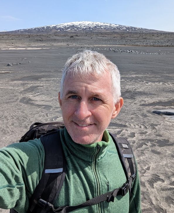 A portrait photo of NASA scientist Ryan Ewing. He wears a backpack and green fleece jacket as he stands in front of an arid tundra landscape.