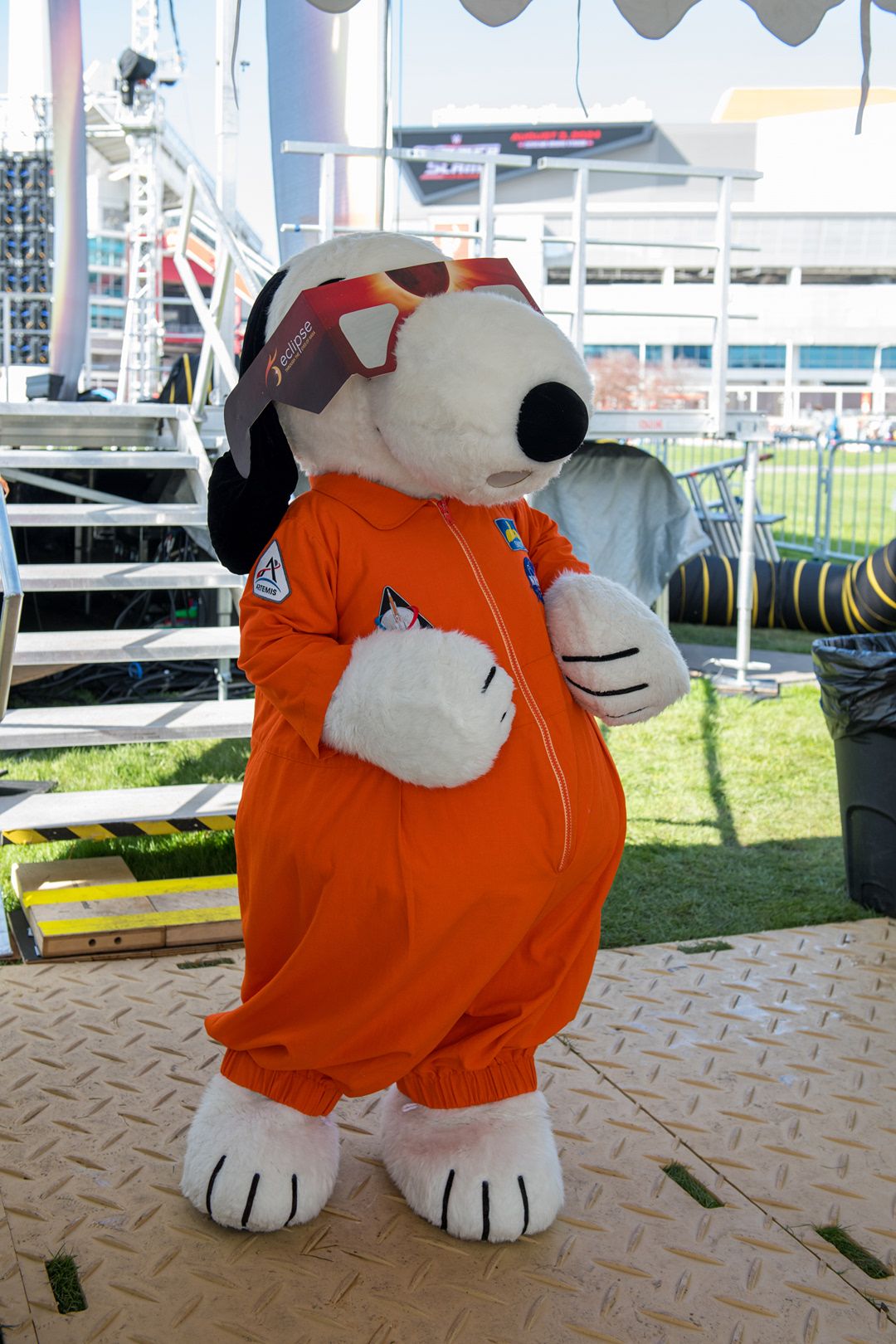 Snoopy, seen as a mascot costume, wearing an orange astronaut suit and large eclipse glasses.