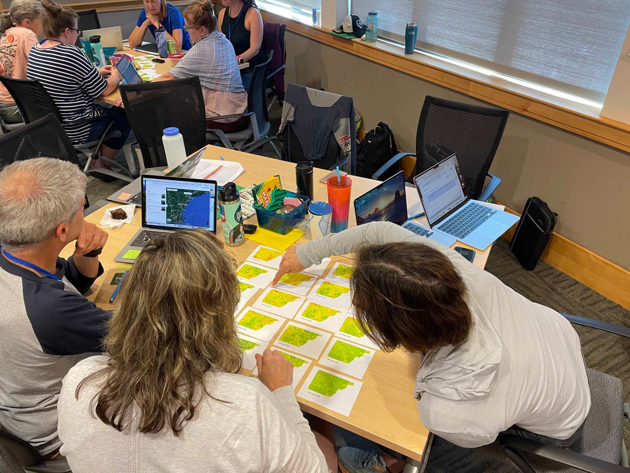 3 teachers at the GMRI summer institute look at a series of NDVI data on small individual maps. One of the teachers is pointing at the small maps, and another had Google Maps open on their nearby computer. In the background, another group of teachers looks at the same data.