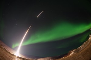 A bright rocket launch streaks across the night sky, leaving a glowing trail of light behind it. In the background, green auroras glow, illuminating the sky with their soft, ethereal light. The scene is set against a mountainous, snow-covered landscape, and the curvature of the ground gives the image a dynamic, wide-angle view. Stars are visible in the clear sky, adding to the sense of vastness and motion.