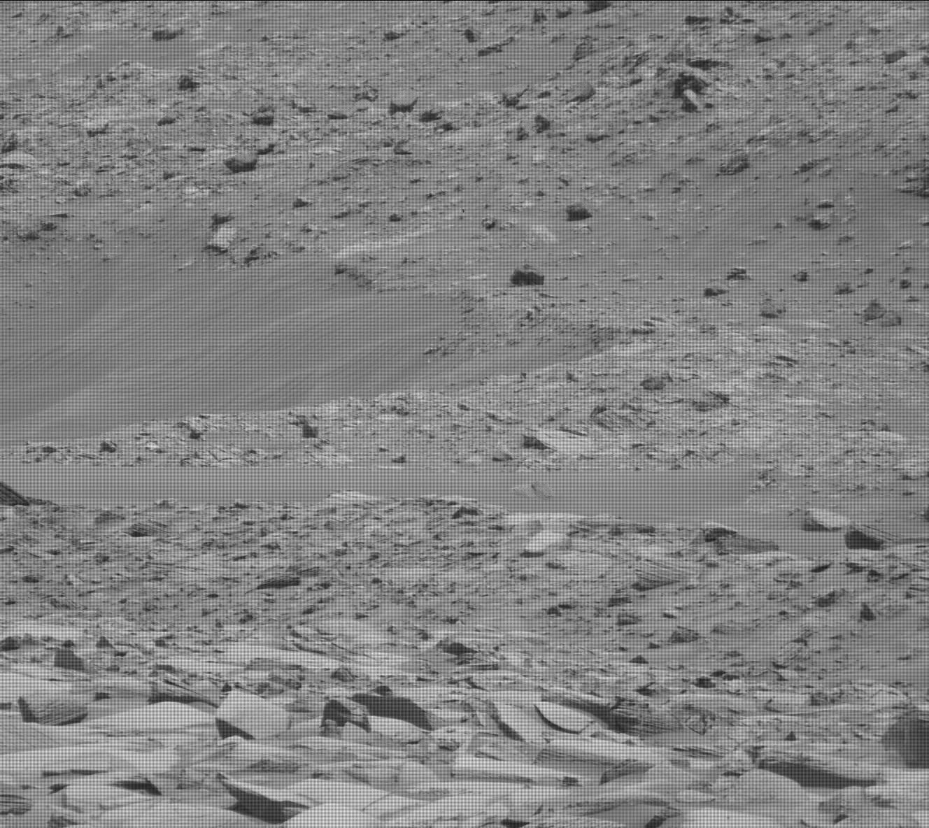 A grayscale image of the Martian surface shows a slightly sloping area of desert-looking rocky terrain in medium gray. An area that looks more smooth and sandy creates a triangle that juts from the left side of the frame and ends at the image center; in the scene it appears to be sloping downward toward the left offscreen. The rest of the terrain is scattered sharp rocks of various sizes.