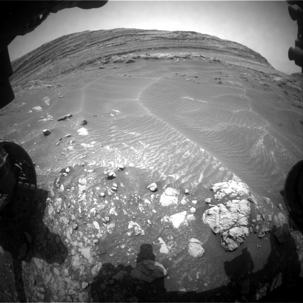 A grayscale, extreme wide-angle, or fish-eye, view of Martian terrain shows a large butte in the distance, on the exaggerated, curved horizon. In the middle of the image, medium gray soil, looking like windblown sand, dominates the scene. In the foreground, amid a shadow of parts of the rover at the bottom of the frame, numerous very bright-toned rocks stand out from the darker surrounding soil.