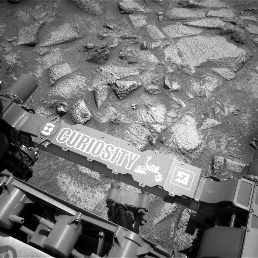 A grayscale photograph of the Martian surface from the Curiosity rover captures medium gray, very uneven terrain in front of the rover, with many angular, medium-sized rocks protruding from the smooth soil. The scene looks like soup with vegetable chunks floating on the surface, or section of brittle candy with scattered nut pieces fused in place. The bottom of the frame shows part of the rover, running from the middle left to the lower right corner of the image, including part of its robotic arm which carries a nameplate imprinted with “Curiosity” outlined in all capital letters, and to the right of that a line drawing of the rover.