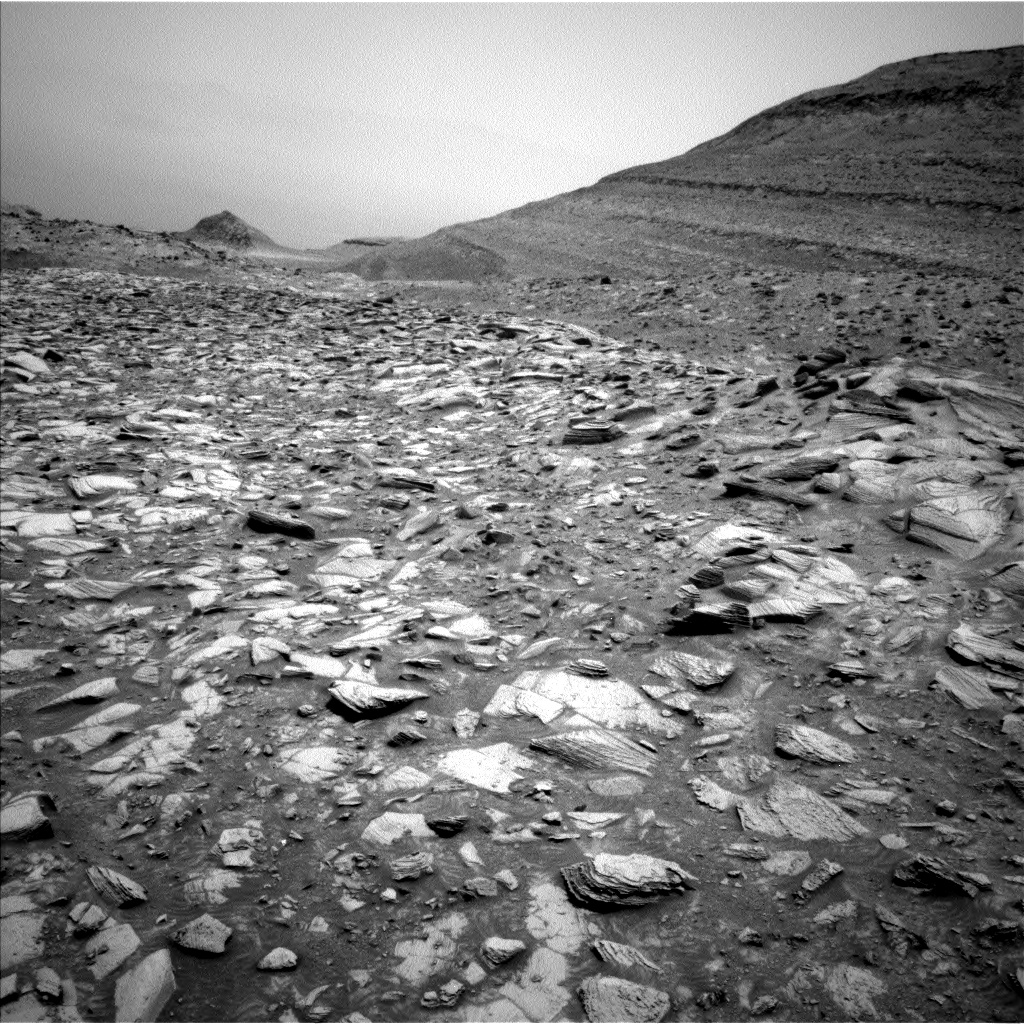 A grayscale photo of the Martian surface shows a long, flat channel extending from the bottom of the frame off into the distance, toward the upper left corner of the image, with medium-gray soil covered by a seemingly endless array of lighter-toned, sharp-edged rocks. Rising from the channel on the right side of the frame is a hill that extends to the upper right corner of the image, which looks smoother, darker, and more striped than the ground below.