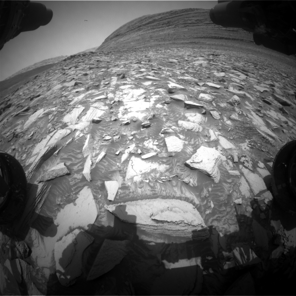 A grayscale photograph from the Martian surface shows a field of very rocky terrain in front of the Curiosity rover, stretching out to a hill rising from the surface, all of it light gray. The field is covered in lighter-toned, medium to large angular rocks. Two of the rover's wheels are visible in the image, at the left and right sides of the frame.