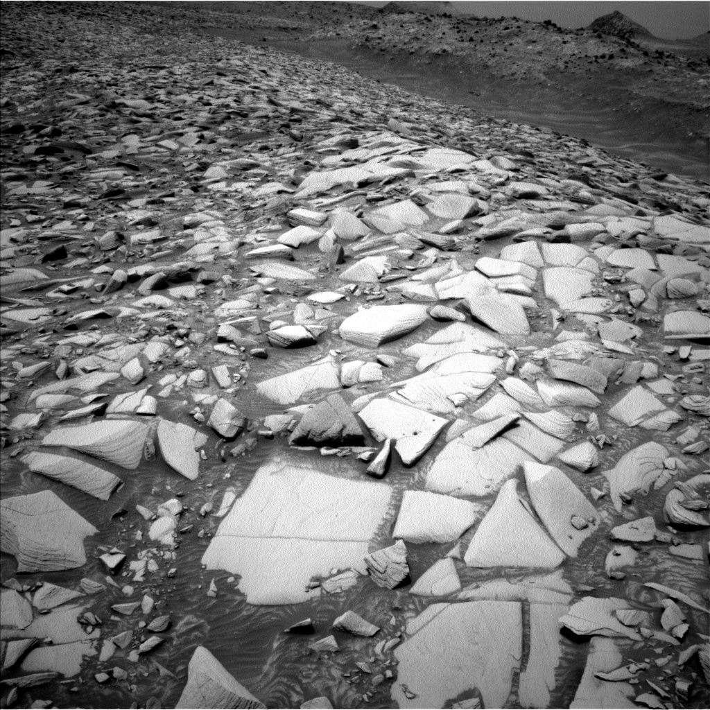 A grayscale photograph of the Martian surface shows a wide expanse of very rocky terrain, with sharp, angular stones arrayed as far as the eye can see, as if set together in a mosaic, stretching to the horizon where distant hills rise up. The stones are very bright and light-toned, while the ground in between them is medium gray, as are the hills and other terrain. A smooth channel cuts through the scene, running diagonally from the top center of the image down to the right side of the frame, just above the middle.