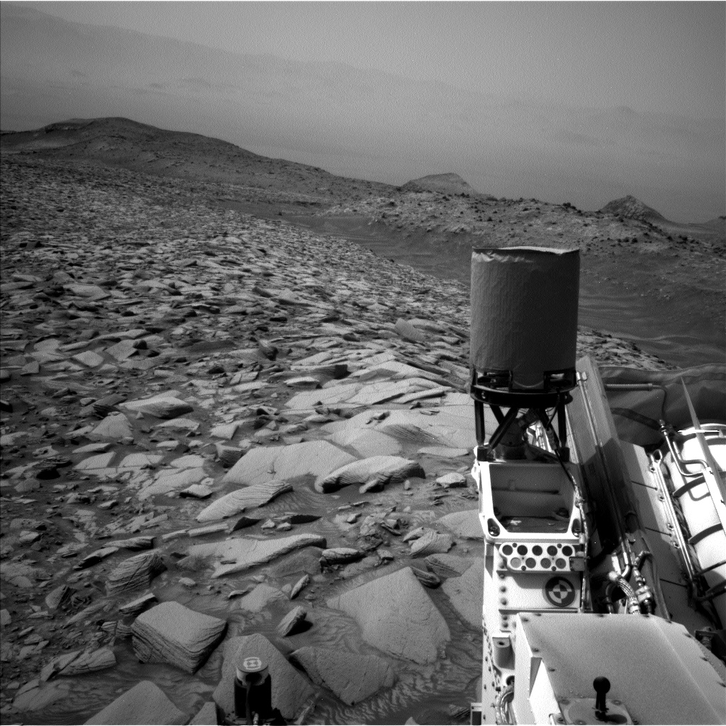 A grayscale photograph of the Martian surface shows a wide expanse of very rocky terrain, with sharp, angular stones arrayed as far as the eye can see, as if set together in a mosaic, stretching to the horizon where distant hills rise up. The stones are very bright and light-toned, while the ground in between them is medium gray, as are the hills and other terrain. A smooth channel cuts through the scene, running diagonally from the top center of the image down to the right side of the frame, just above the middle. A portion of the Curiosity rover is visible in the lower-right corner of the frame.