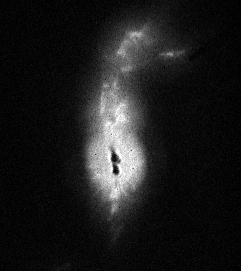 Black and white image of a bright-white cloud. It is orientated vertically and holds many bright knots and rays. Two black, ink-blot splotches sit vertically at image center