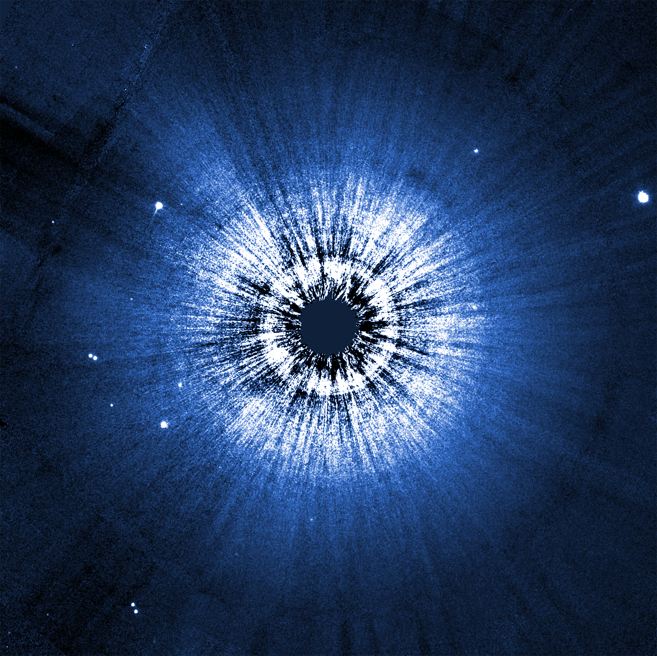 A Hubble image of the circumstellar disk around Vega. The disk is perfectly circular, and at the center is a black spot blocking out the bright glow of a star. Closer to the center, the disk is white. Radial striations extend out from the center, giving a ripple effect to the disk like the end of a sausage casing. The outer edge of the circular disk is blue.