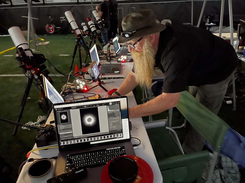 There is a long table with laptops showing views of the total solar eclipse. A person leans over one computer. Lined up against the table are several telescopes pointing up.