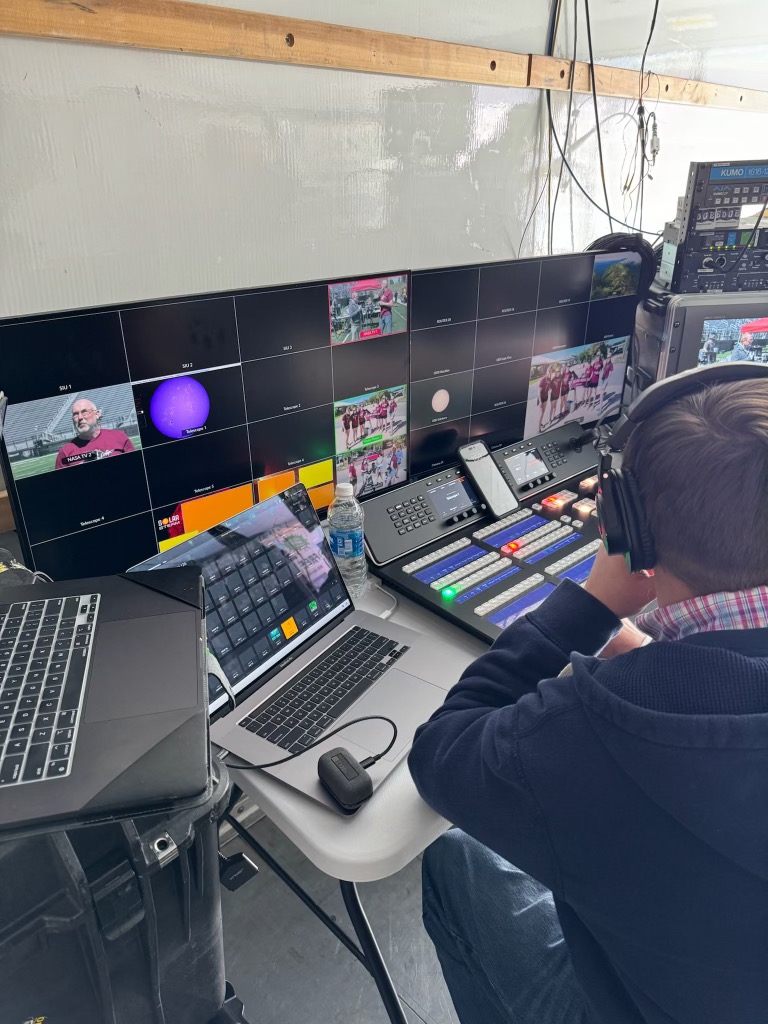 A man sits at a table filled with computers and screens, showing views from the broadcast.