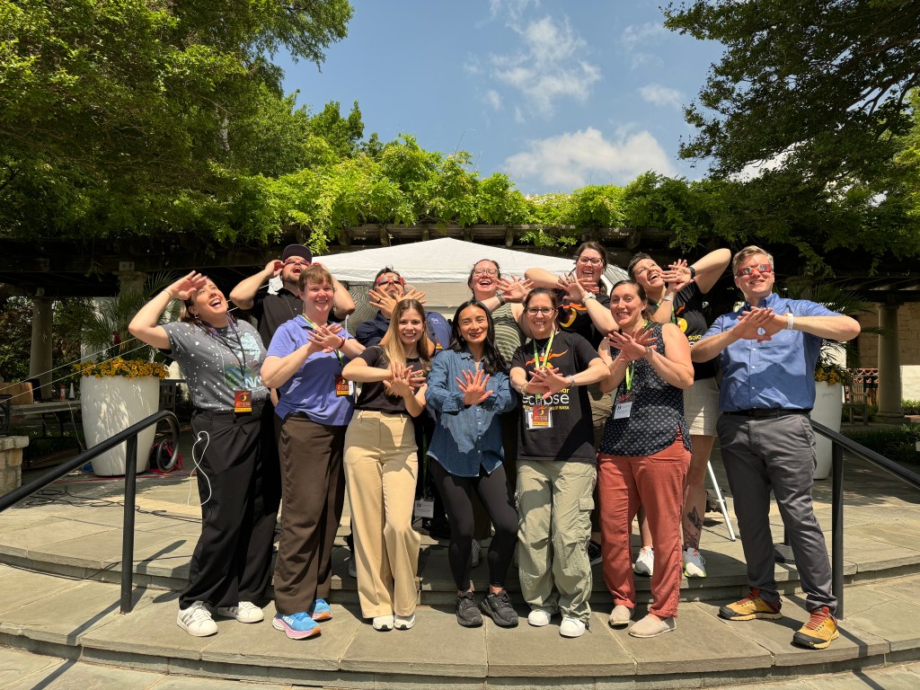 A group of people stand outside. Many of them are crossing their hands in front of each other, with their fingers spread out. Some are wearing eclipse glasses.