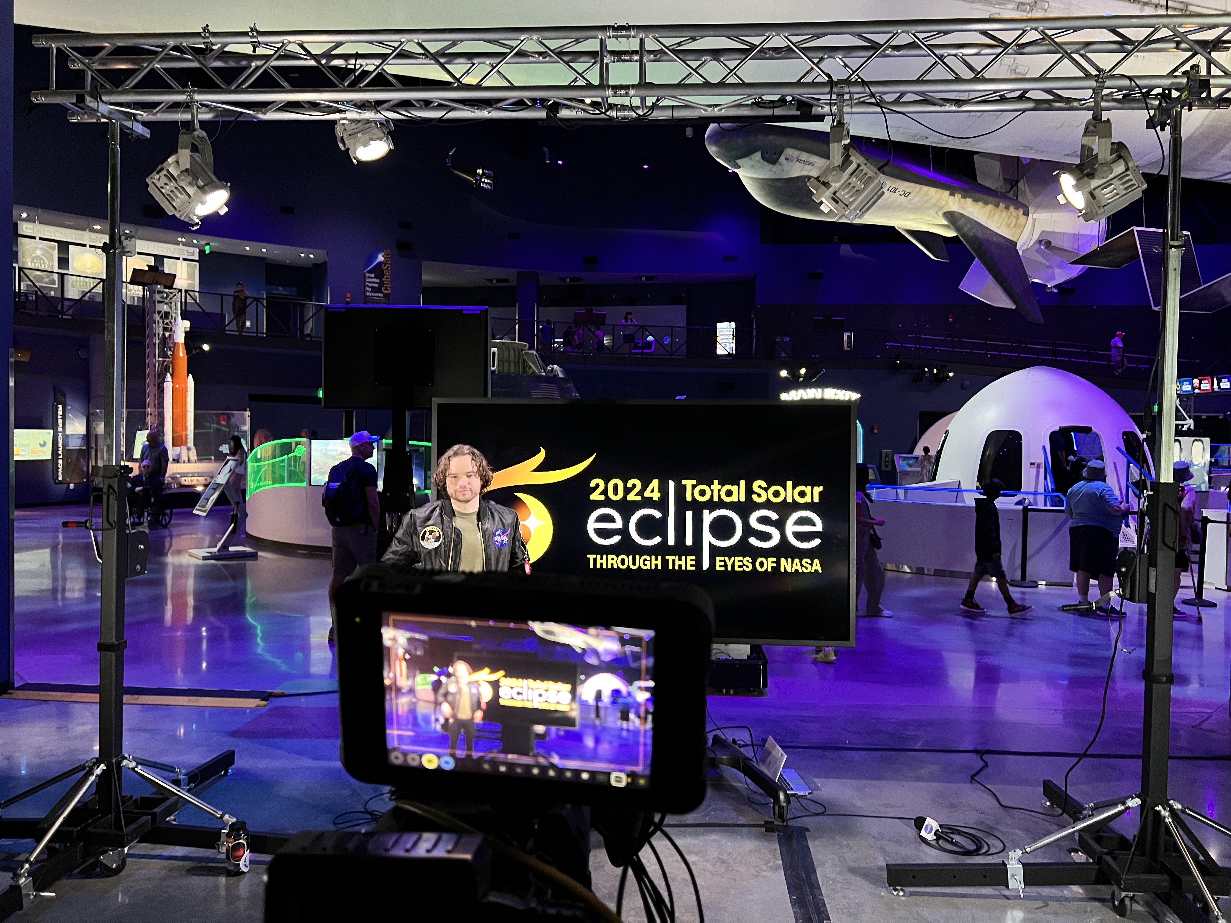 A man stands in front of a screen that says Eclipse Through the Eyes of NASA. Behind him is a museum, with space themed items and a space craft hanging from the ceiling.