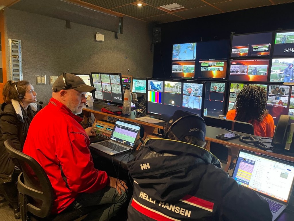 Inside a production truck, multiple people sit in front of computers with large screens showing several views in front of them.