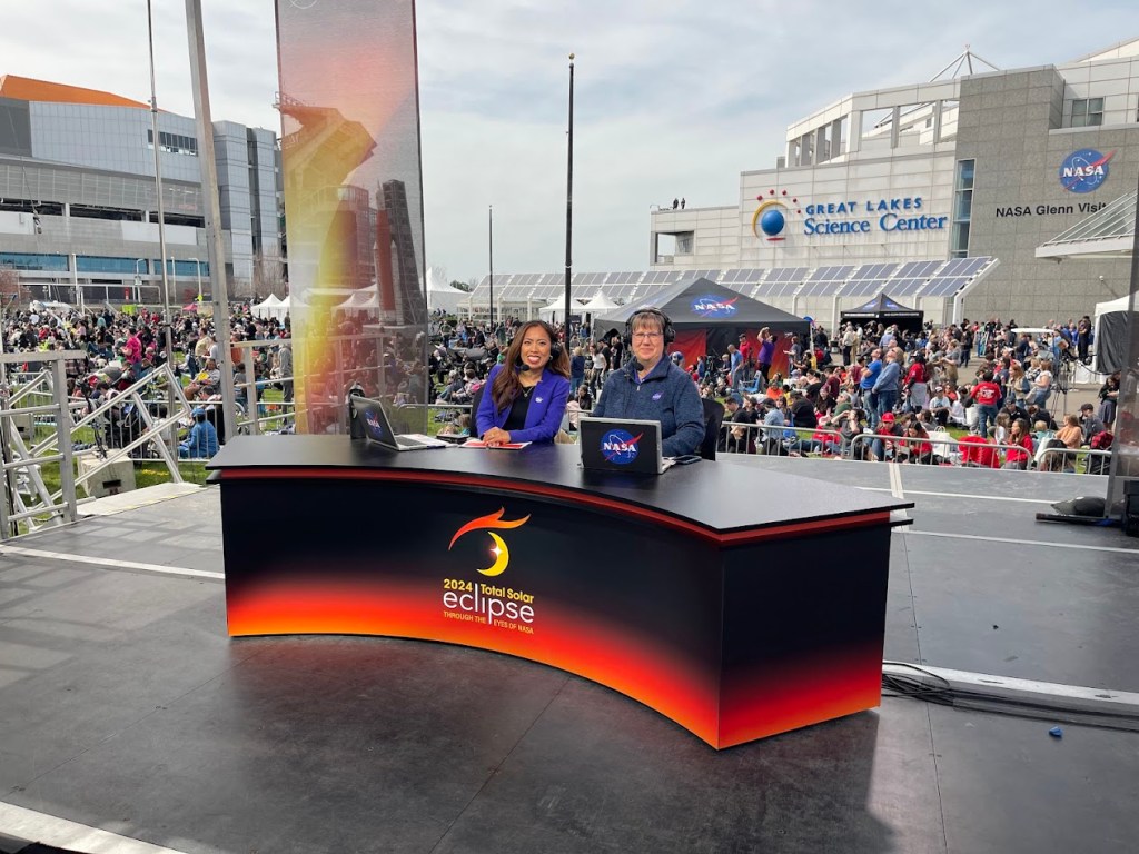 NASA eclipse broadcast host Megan Cruz and co-host Sarah Noble sit on stage in front of the Great Lakes Science Center in Cleveland, OH.