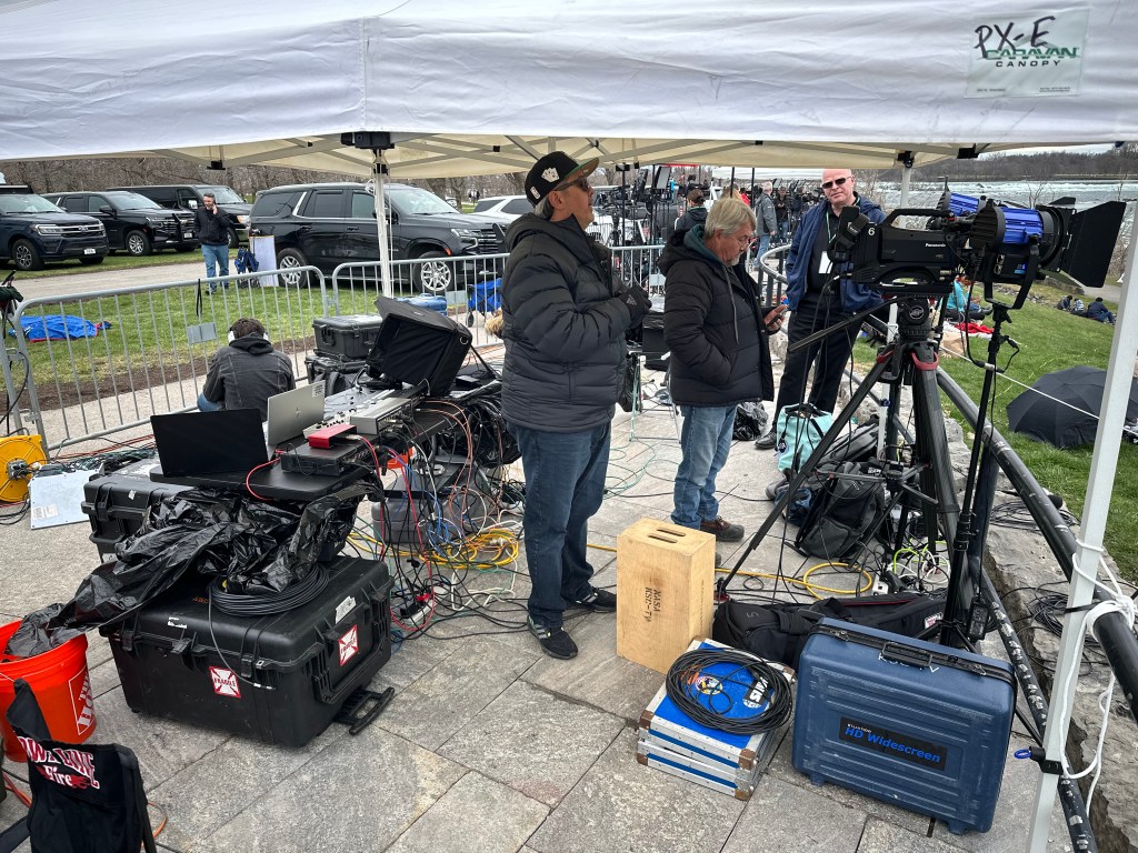 Two people stand under a tent surrounded by cameras and other broadcast equipment.