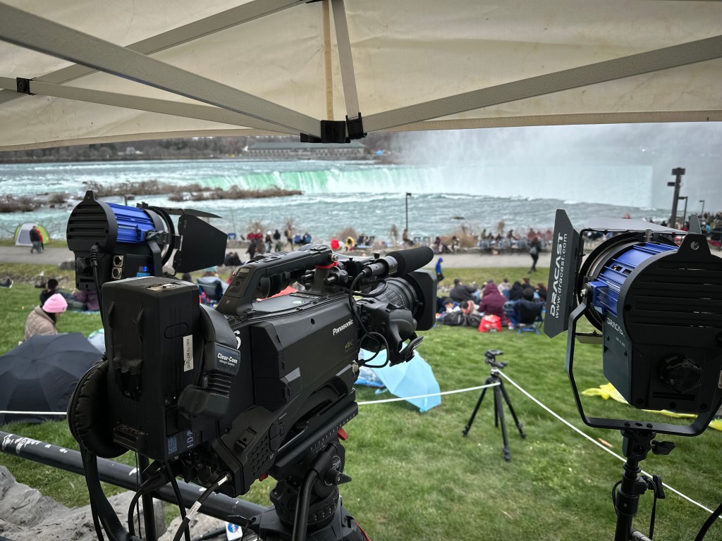 Camera in front of a crowd on grass, which is looking out toward a large body of water.