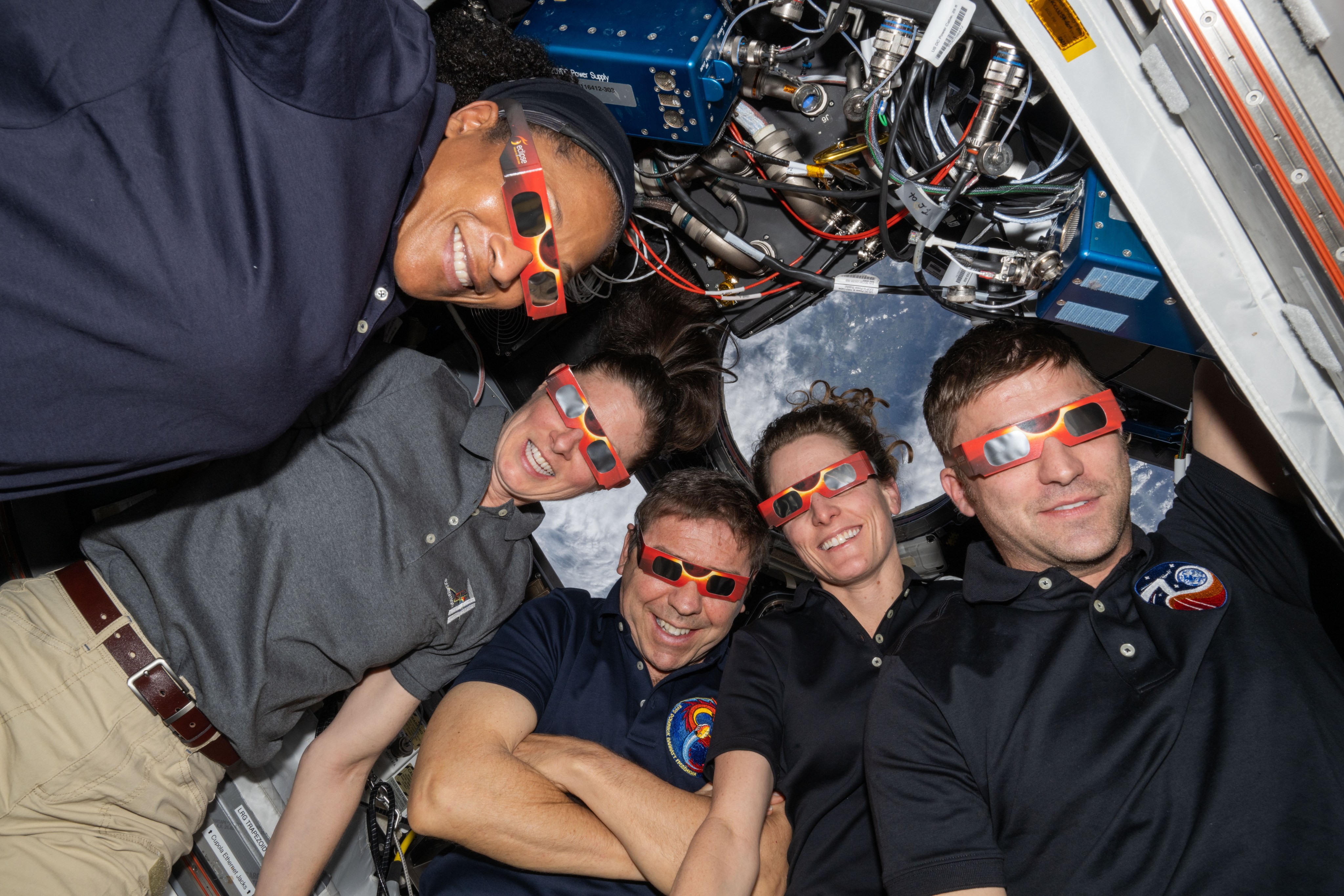 Five astronauts aboard the International Space Station wear red eclipse glasses. Behind them, you can see Earth.