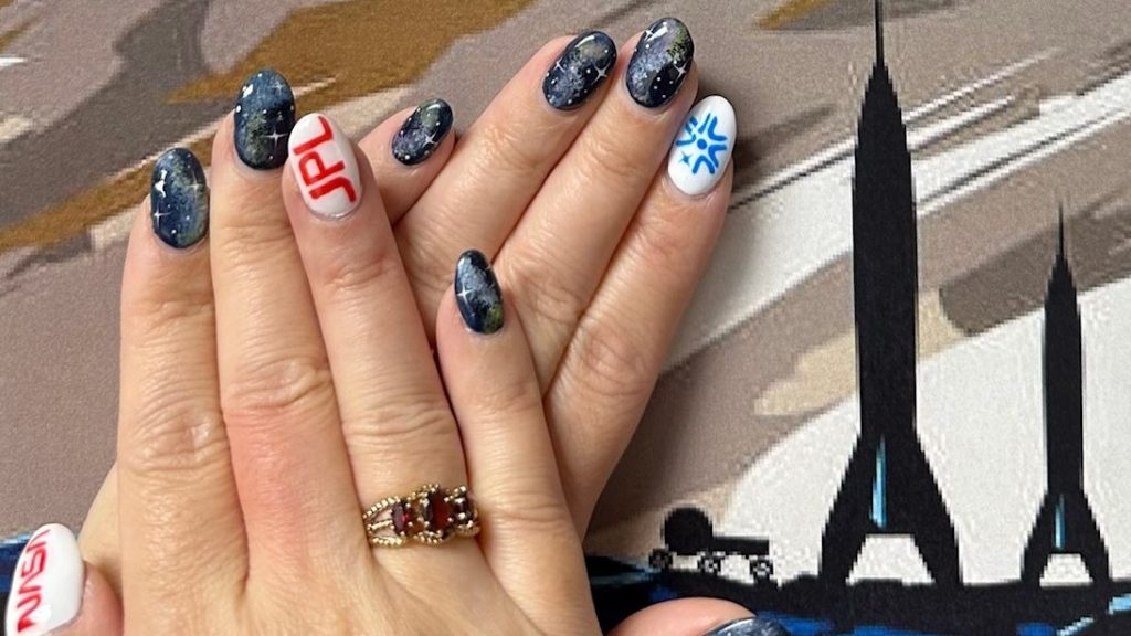 Fingernails painted with colors symbolic of the Europa Clipper mission. The hands are next to art with rockets.