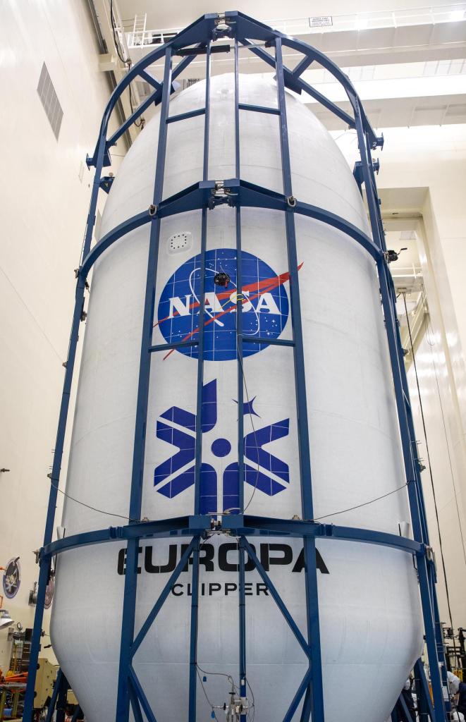 Europa Clipper's white payload fairing covers the vehicle. The NASA logo and blue mission identifier are painted on the front. A blue structure holds the spacecraft upright.