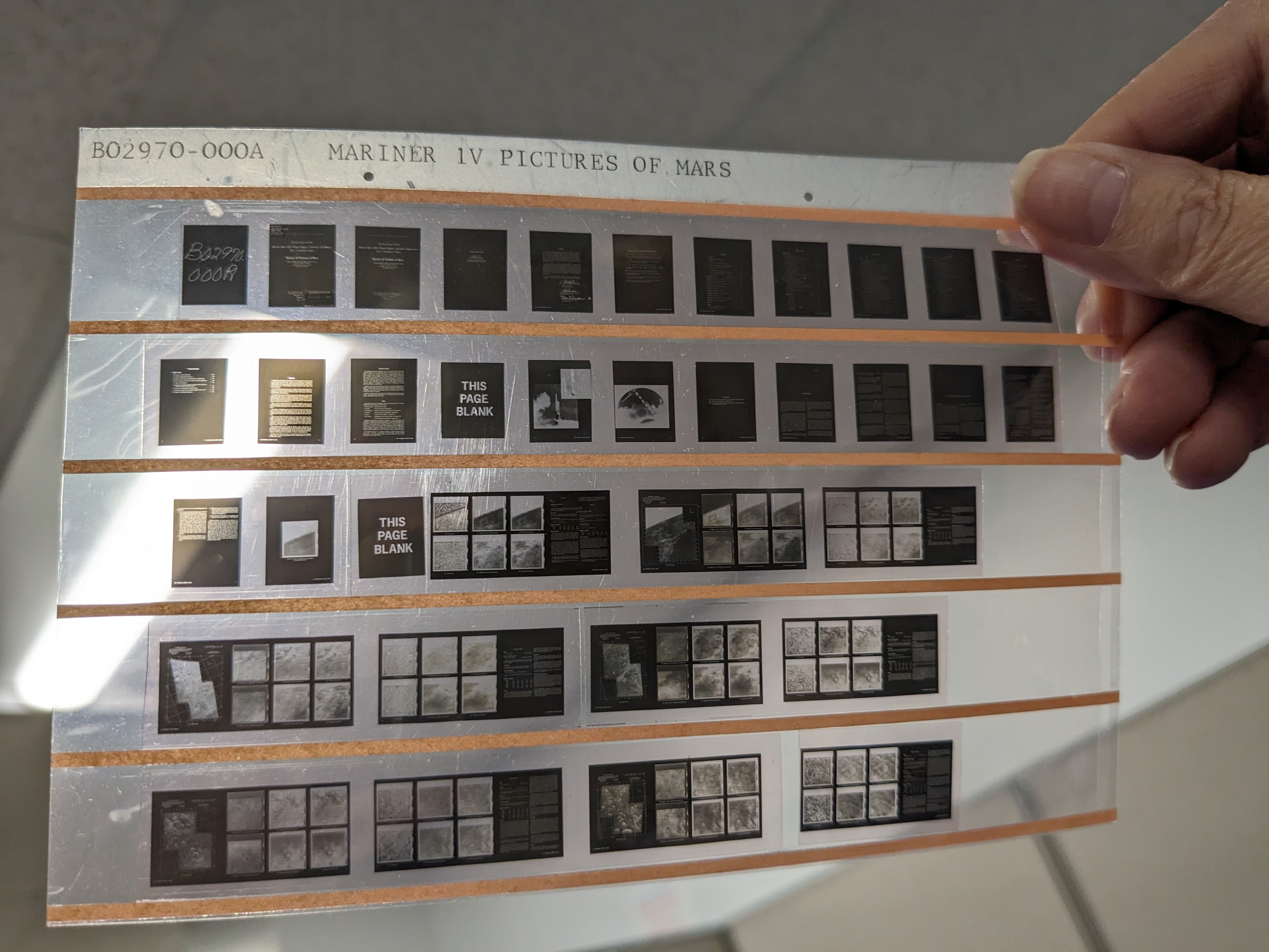 Three side-by-side images. First Image: A collection of rectangular boxes stacked in two rows. Each box has a white label with text detailing what’s inside, including experiments and film types. Some labels have handwritten notes and dates. The boxes are neatly arranged on a shelf. Second Image: A hand holding a strip of microfilm up to the light. The strip contains several rows of tiny black-and-white images and text blocks. The top of the strip reads "Mariner IV Pictures of Mars," and some frames are blank, while others show faint images or text. Third Image: A black-and-white image of Jupiter on a screen. The planet's cloudy bands are visible, with a small white spot near the center. A gloved hand points at the image, which is framed by a screen with data and numbers at the bottom.