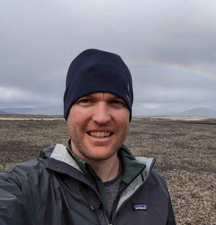 A portrait photo of NASA scientist Matthew Miller outdoors. He wears a gray Patagonia jacket and a blue beanie in front of an arid landscape.