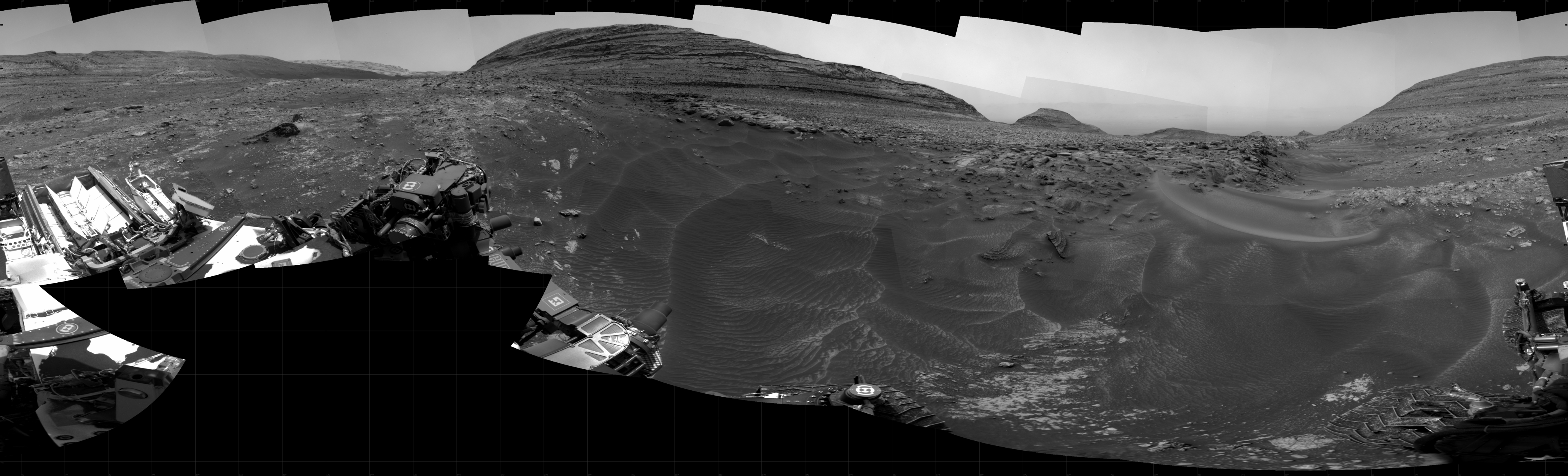 A grayscale panorama of the Martian surface shows a wide field of flat terrain dotted with small rocks, all in dark gray, stretching into the distance where a large butte rises from the ground. A portion of the Curiosity rover is visible in the lower left corner of the image.