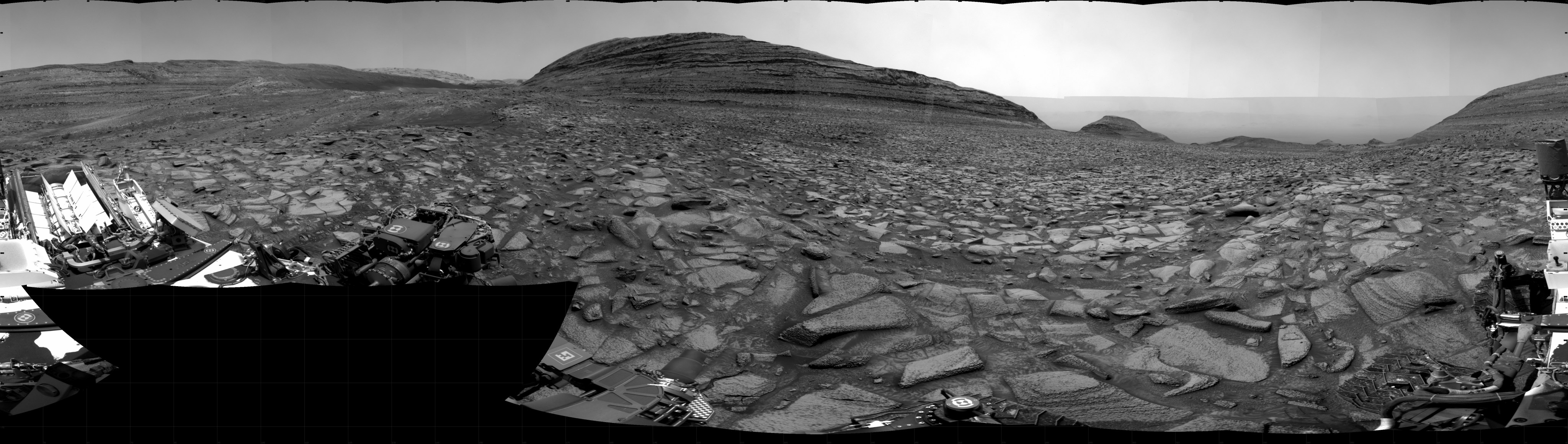 A grayscale panorama of the Martian surface shows a brightly lit, wide field of flat terrain dotted with flat, angular, medium rocks, all in dark gray, stretching into the distance where a large butte rises from the ground. A portion of the Curiosity rover is visible in the lower left and lower right corners of the image.