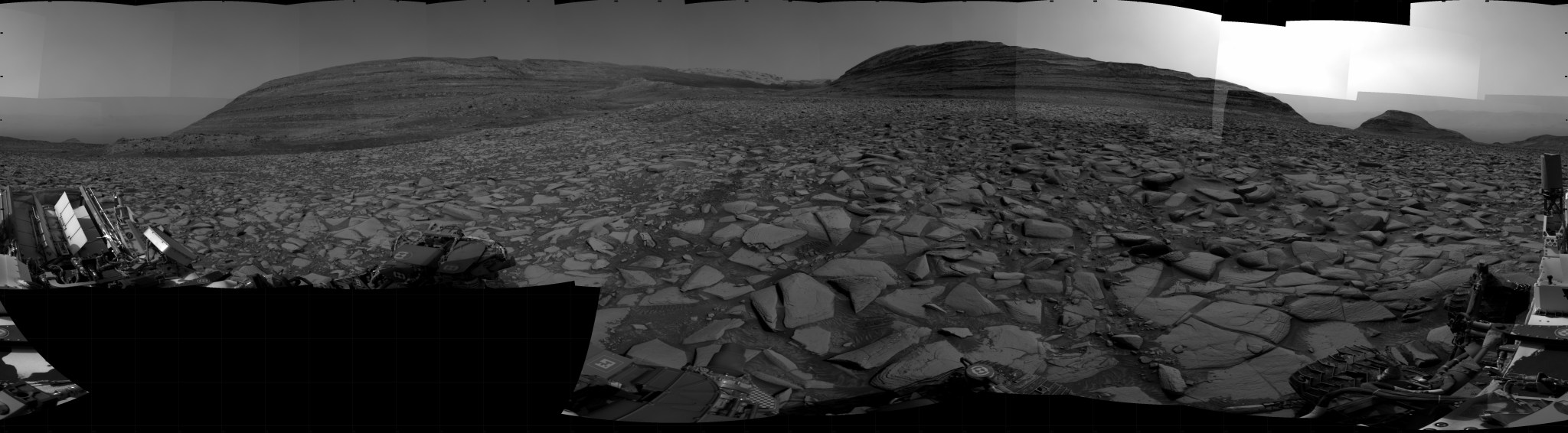 A grayscale panorama of the Martian surface shows a wide field of flat terrain dotted with flat, angular, medium-sized rocks, all in dark gray, stretching into the distance where a series of hills rise from the ground. A portion of the Curiosity rover is visible at the left and right sides of the image.