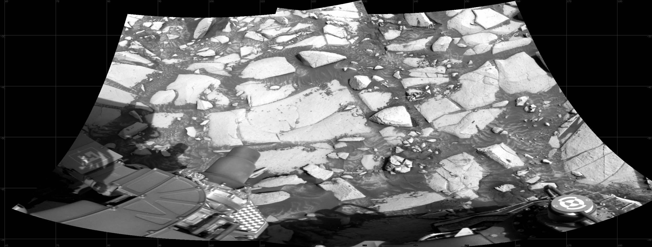 A trapezoid-shaped grayscale image from the Martian surface shows medium gray soil covered in bright-toned, almost whitish rocks of all shapes and sizes, filling the frame like parts of a mosaic. A portion of the Curiosity rover is visible at the bottom and lower left corner of the frame.