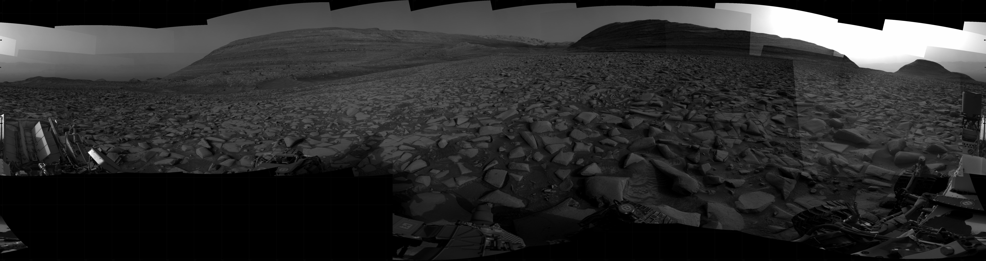 A grayscale panoramic image of the Martian surface shows dark skies and ground, where the rolling terrain is marked by uneven slabs of rock covered in some areas by soil and small rocks. In the background, gently sloped, layered rock hills rise above the ground at top left and right. The hill on the right is hiding the Sun, which is illuminating the only bright area in the picture, the sky above and to the sides of the hill. Portions of the Curiosity rover are visible at the left, right, and bottom edges of the extreme wide-angle image.