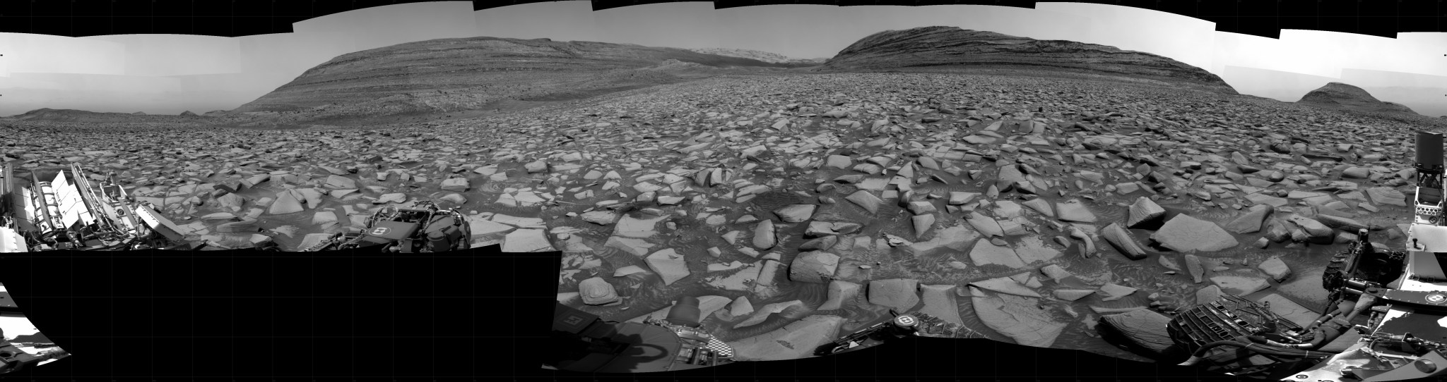 A grayscale panorama of the Martian surface shows a wide field of flat terrain dotted with flat, angular, medium-sized rocks, all in dark gray, stretching into the distance where a series of hills rise from the ground. A portion of the Curiosity rover is visible at the left and right sides of the image.