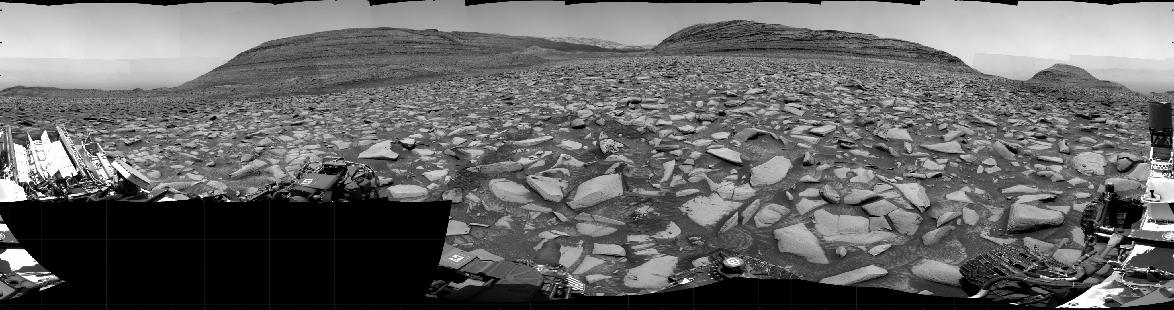 A grayscale panorama of the Martian surface shows a wide field of flat terrain dotted with flat, angular, medium-sized rocks, all in dark gray, stretching into the distance where a series of hills rise from the ground. A portion of the Curiosity rover is visible at the left and right sides of the image.