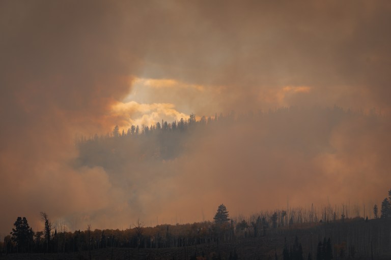 A photograph of a smoky landscape. Most of the image is shades of brown, with nothing visible through the smoke. Near the center of the image the air is somewhat clearer, showing a window-like view of a mountainous landscape with blue skies above.A photograph of a smoky landscape. Most of the image is shades of brown, with nothing visible through the smoke. Near the center of the image the air is somewhat clearer, showing a window-like view of a mountainous landscape with blue skies above.