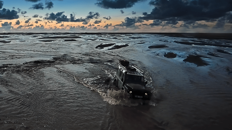 An aerial shot of a green, off-road jeep drives through a flooded plain, sending water into the air as it drives across an Icelandic landscape.