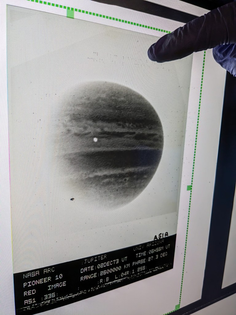 Three side-by-side images. First Image: A collection of rectangular boxes stacked in two rows. Each box has a white label with text detailing what’s inside, including experiments and film types. Some labels have handwritten notes and dates. The boxes are neatly arranged on a shelf. Second Image: A hand holding a strip of microfilm up to the light. The strip contains several rows of tiny black-and-white images and text blocks. The top of the strip reads 