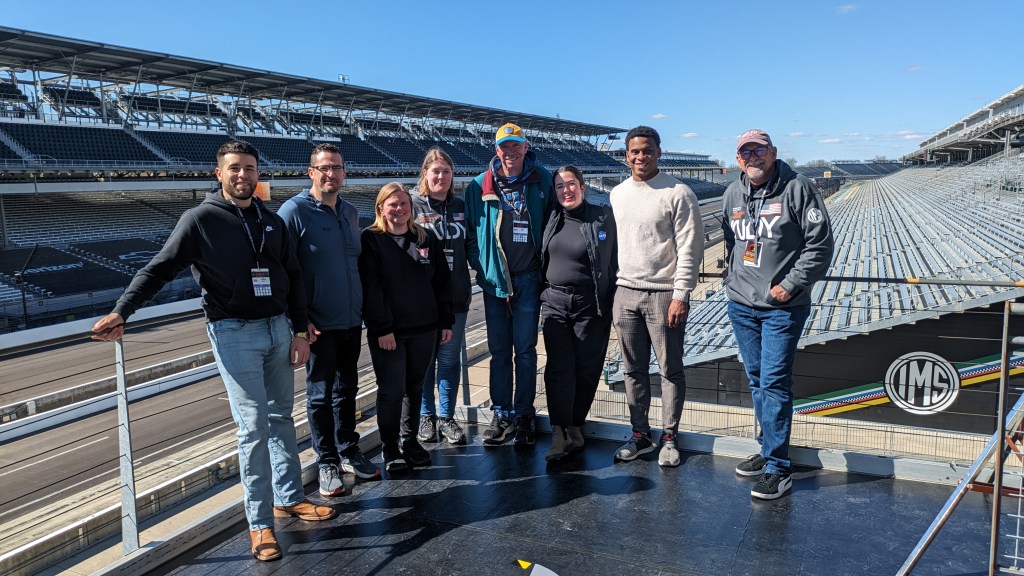 The team supporting the NASA broadcast from Indianapolis Motor Speedway in Indianapolis pose for a photo with the racetrack in the background.