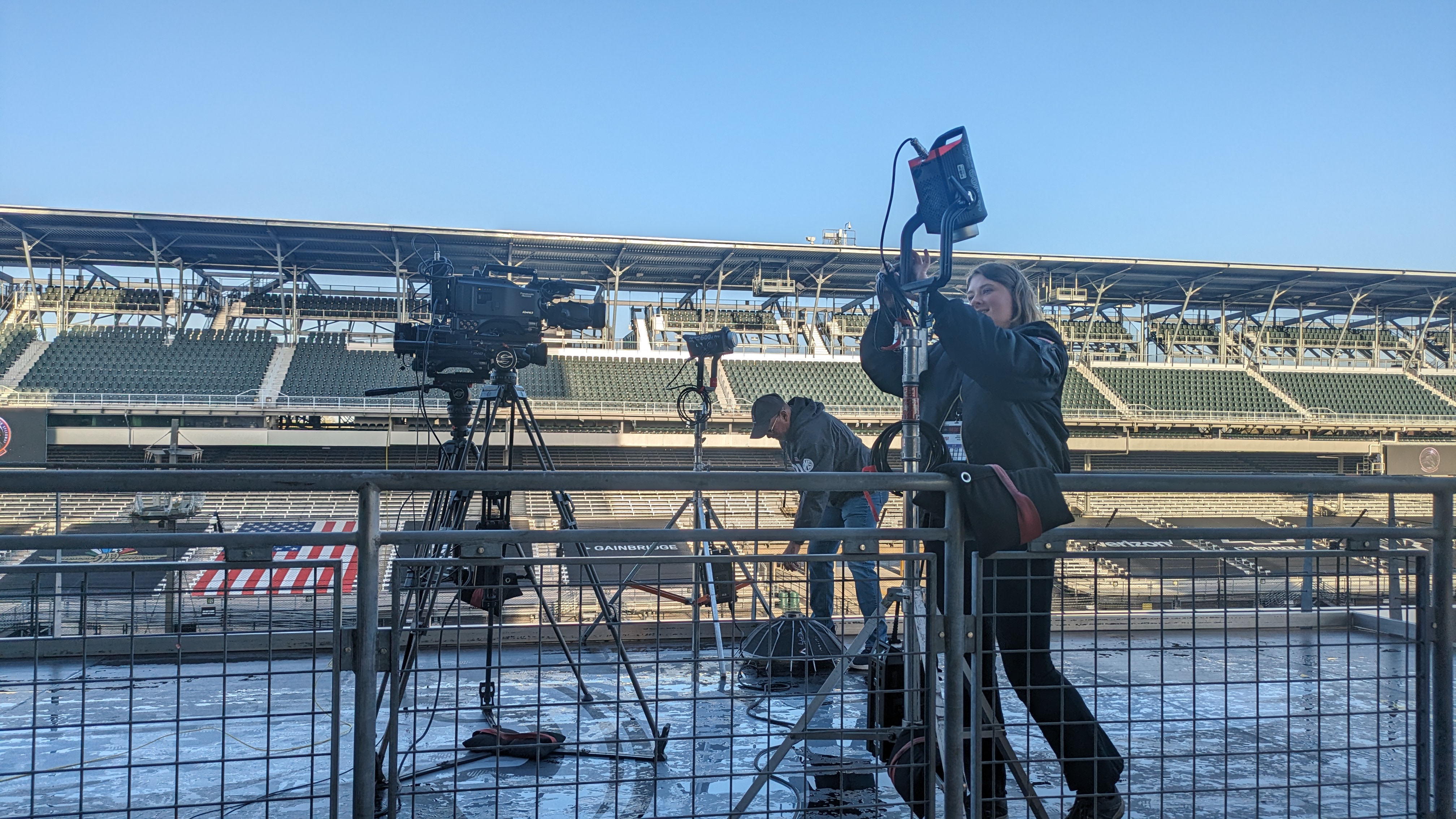 The crew in Indianapolis, IN set up cameras the broadcast location at the Indianapolis Motor Speedway. The racetrack and stands can be seen in the background.