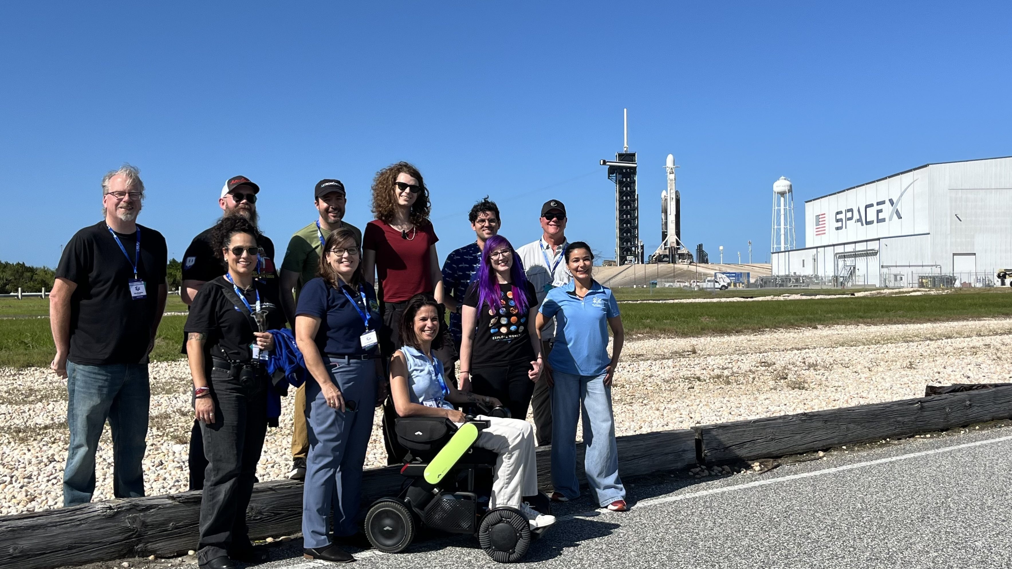 Eleven people gather on a road near launch pad 39A at Kennedy Space Center. Europa Clipper can be seen behind them, and the right of them is the SpaceX hangar.