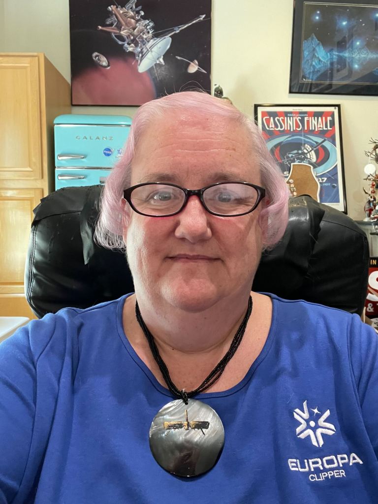 A woman wearing glasses, a blue shirt with the Europa Clipper Mission Identifier, and silver necklace with a spacecraft on it.