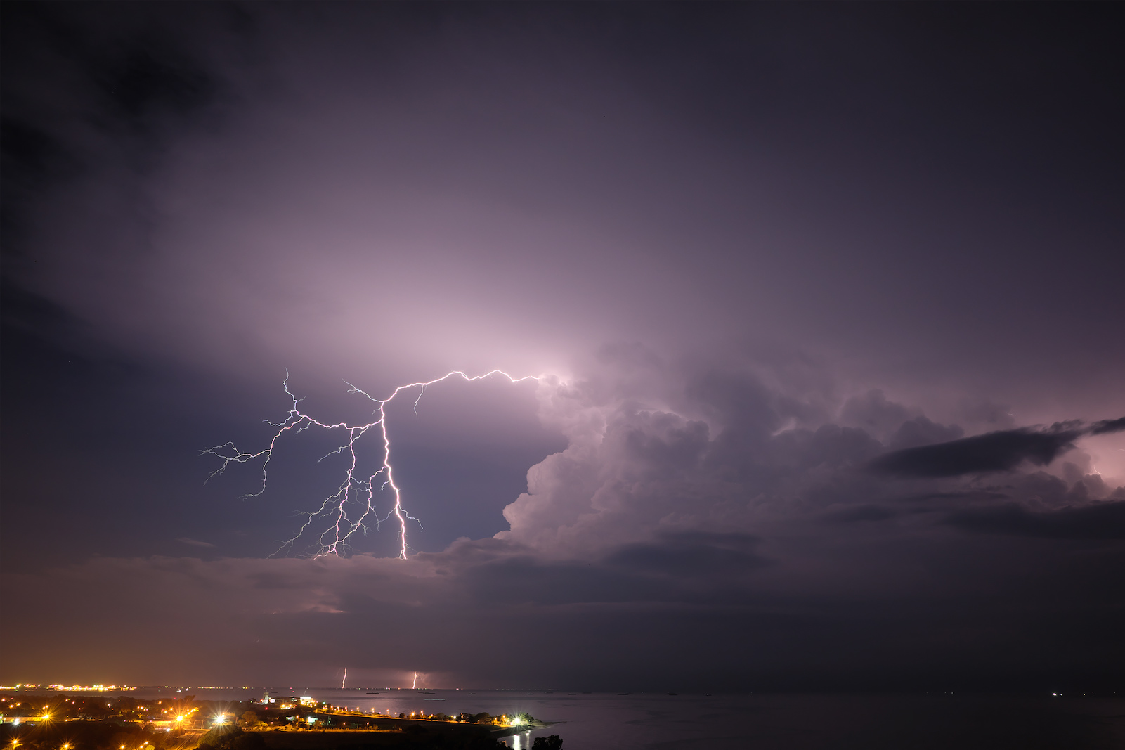 Two lightning strikes are visible in the distance with city lights along the left of the image along the coastline of the bay that occupies the lower right and curves into the