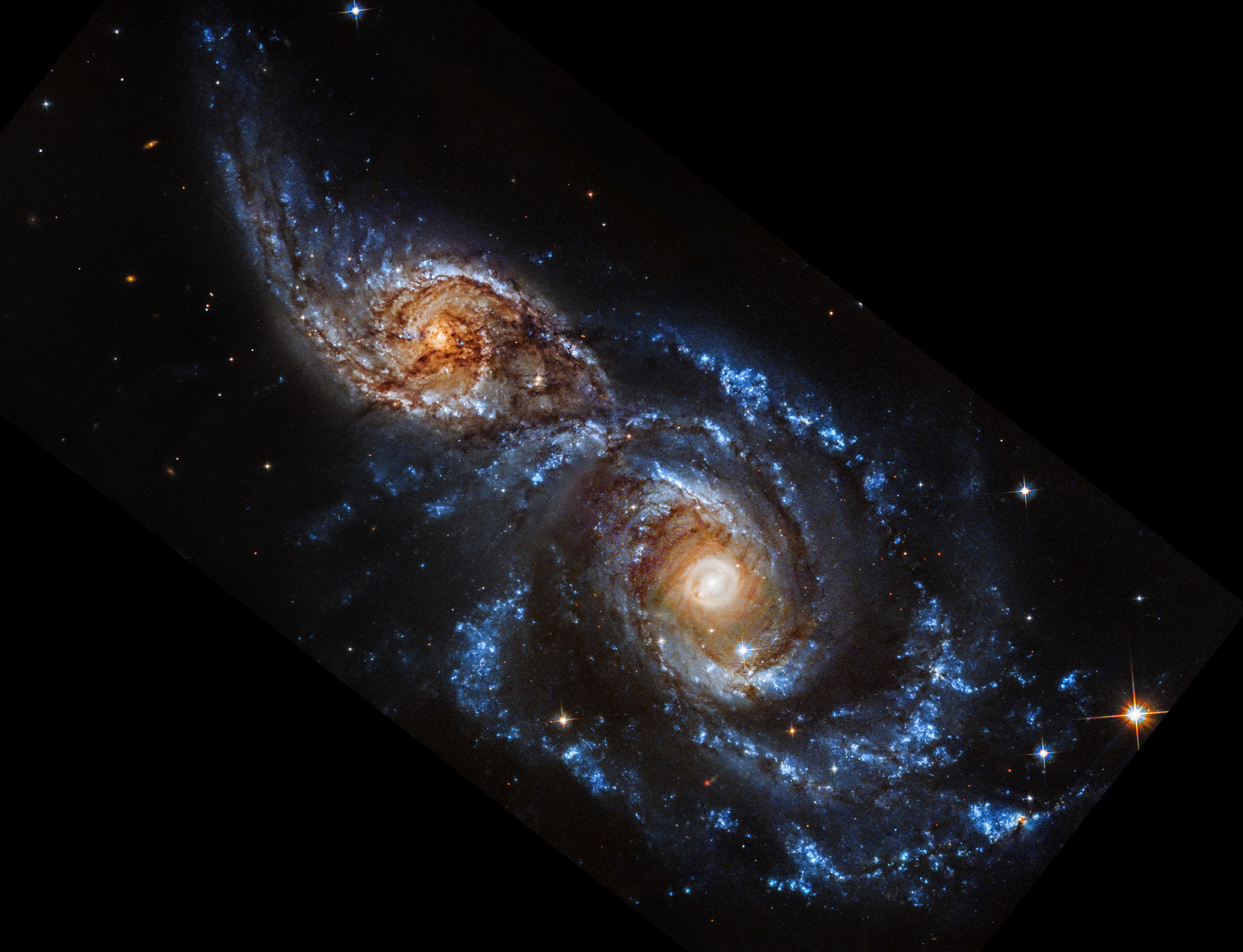 The Hubble View cut from two views of the same object shown side by side.. The Hubble observation is at left, and the Webb observation is at right. Both show an angled pair of spiral galaxies, IC 2163 at top left, and NGC 2207, at bottom right. IC 2163 is smaller than NGC 2207, and they appear to be overlapping at the center diagonal. In the Hubble image, the galaxies’ cores appear white at the center, and more orange overall. Both galaxies have bright blue spiral arms, and are interlaced with dark brown, irregular dust lanes. Tiny galaxies and a few foreground stars dot the black background of space. There are no data along the bottom left, top right, and bottom right corners. In the Webb image, the galaxies’ cores appear bright white, transitioning to spiral arms that are also bright white, but flecked with pink dots. Tiny galaxies and a few foreground stars dot the entire black background of space.