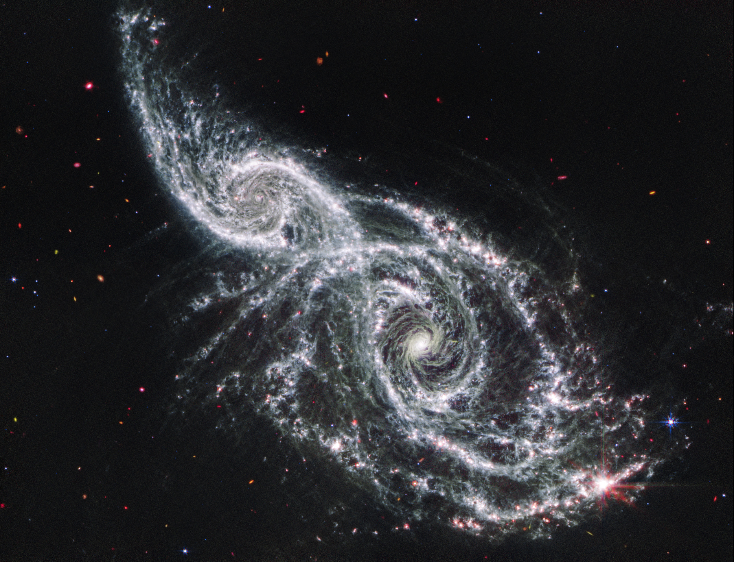 The Webb View cut from two views of the same object shown side by side.. The Hubble observation is at left, and the Webb observation is at right. Both show an angled pair of spiral galaxies, IC 2163 at top left, and NGC 2207, at bottom right. IC 2163 is smaller than NGC 2207, and they appear to be overlapping at the center diagonal. In the Hubble image, the galaxies’ cores appear white at the center, and more orange overall. Both galaxies have bright blue spiral arms, and are interlaced with dark brown, irregular dust lanes. Tiny galaxies and a few foreground stars dot the black background of space. There are no data along the bottom left, top right, and bottom right corners. In the Webb image, the galaxies’ cores appear bright white, transitioning to spiral arms that are also bright white, but flecked with pink dots. Tiny galaxies and a few foreground stars dot the entire black background of space.