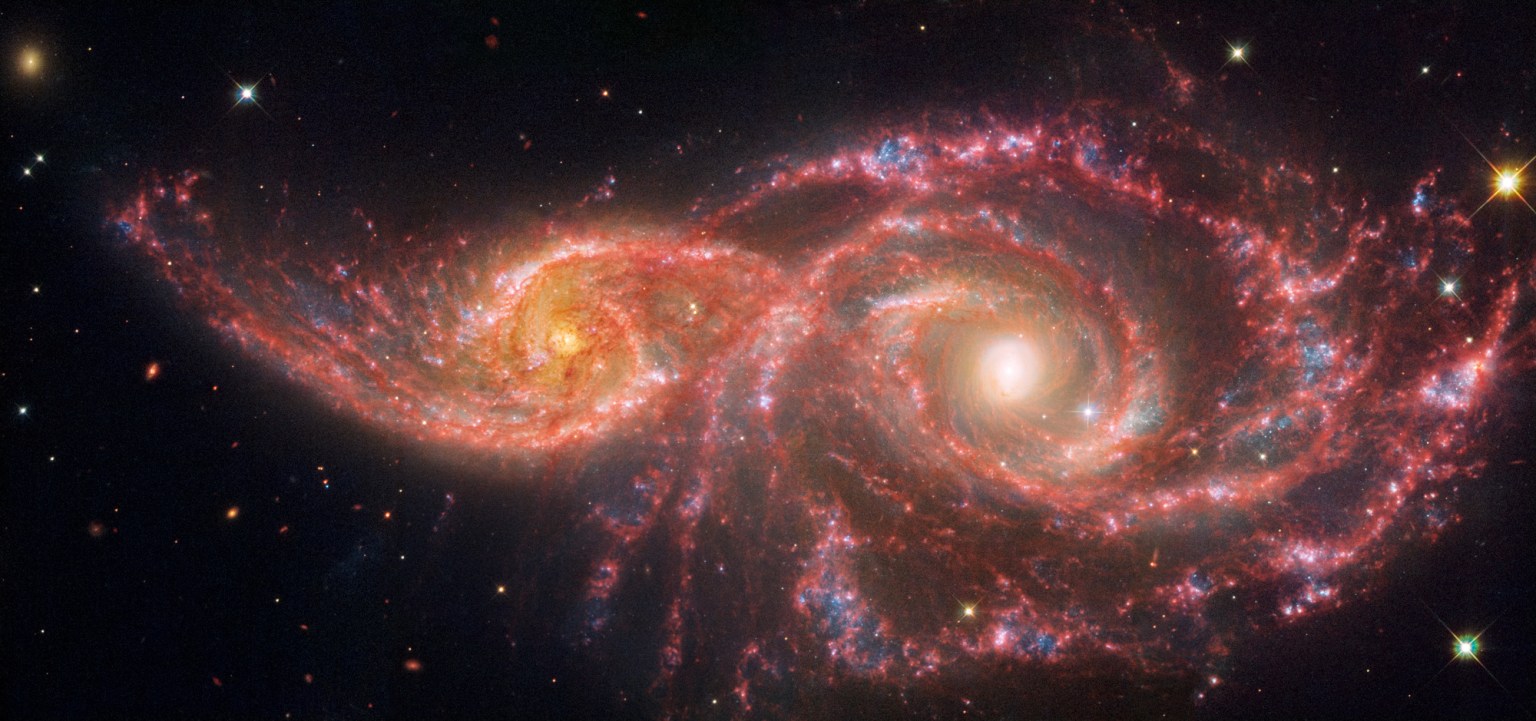 Two spiral galaxies take the shape of a colorful beaded mask that sits above the nose. The galaxy at left, IC 2163, is smaller, taking up a little over a quarter of the view. The galaxy at right, NGC 2207, takes up half the view, with its spiral arms reaching the edges. IC 2163 has a bright orange core, with two prominent spiral arms that rotate counter clockwise and become straighter towards the ends, the left side extending almost to the edge. Its arms are a mix of pink, white, and blue, with an area that takes the shape of an eyelid appearing whitest. NGC 2207 has a very bright core. Overall, it appears to have larger, thicker spiral arms that spin counter clockwise. This galaxy also contains more and larger blue areas of star formation that poke out like holes from the pink spiral arms. In the middle, the galaxies' arms appear to overlap. The edges show the black background of space, including extremely distant galaxies that look like orange and red smudges, and a few foreground stars.