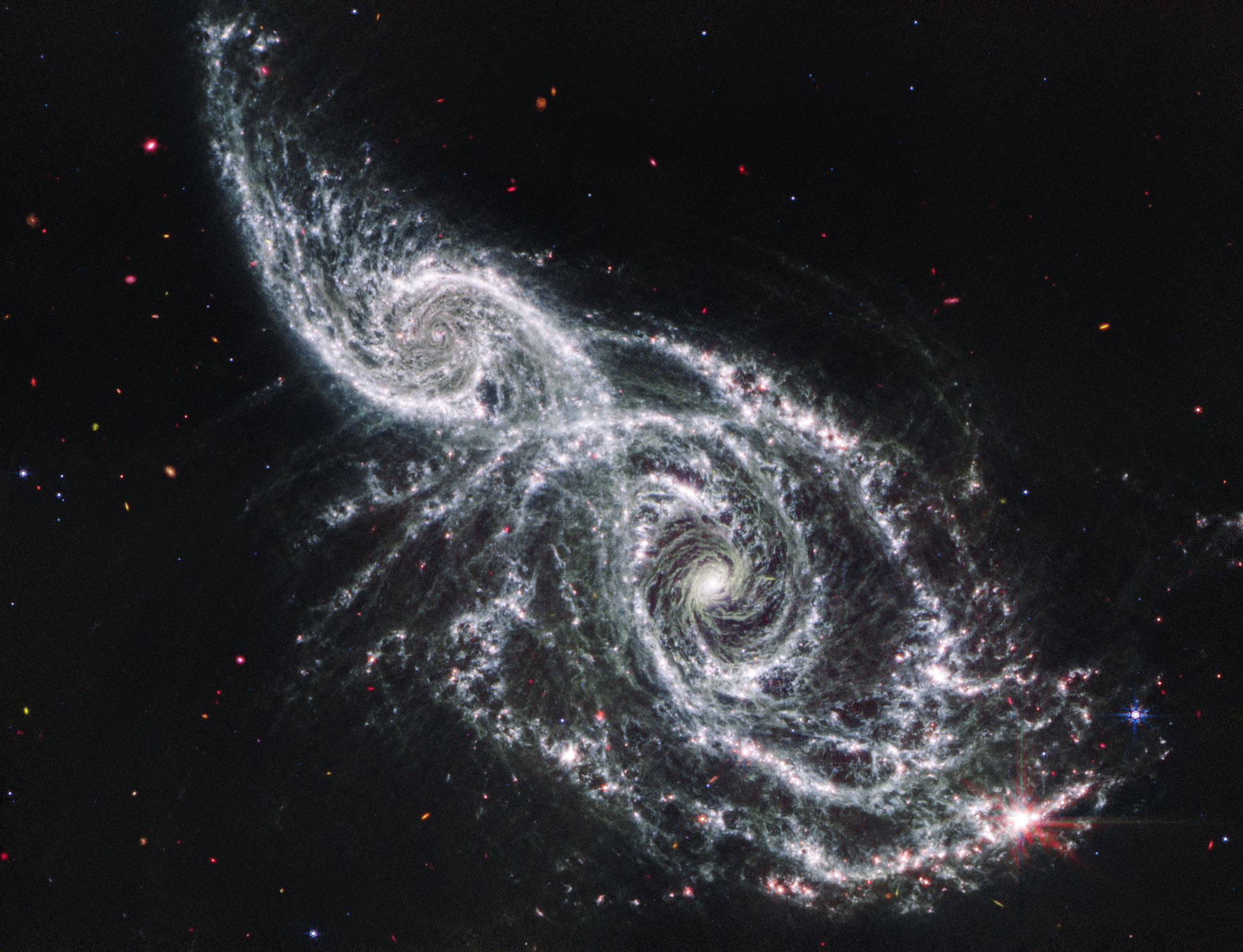 Two spiral galaxies take the shape of a colorful beaded mask that sits above the nose and is angled from top left to bottom right. The galaxy at left, IC 2163, is smaller, taking up a little over a quarter of the view. The galaxy at right, NGC 2207, takes up half the view. IC 2163 has a bright white core, with two prominent white spiral arms that rotate counter clockwise and become straighter toward the ends. Its arms are dotted with pink. NGC 2207 has a very bright core that is larger. Overall, it appears to have thicker spiral arms that spin counter clockwise. This galaxy also contains more pink areas of star formation. In the middle, the galaxies’ arms appear to overlap. The black background of space shows extremely distant galaxies that are tiny, and often red and orange smudges, as well as a few blue foreground stars.