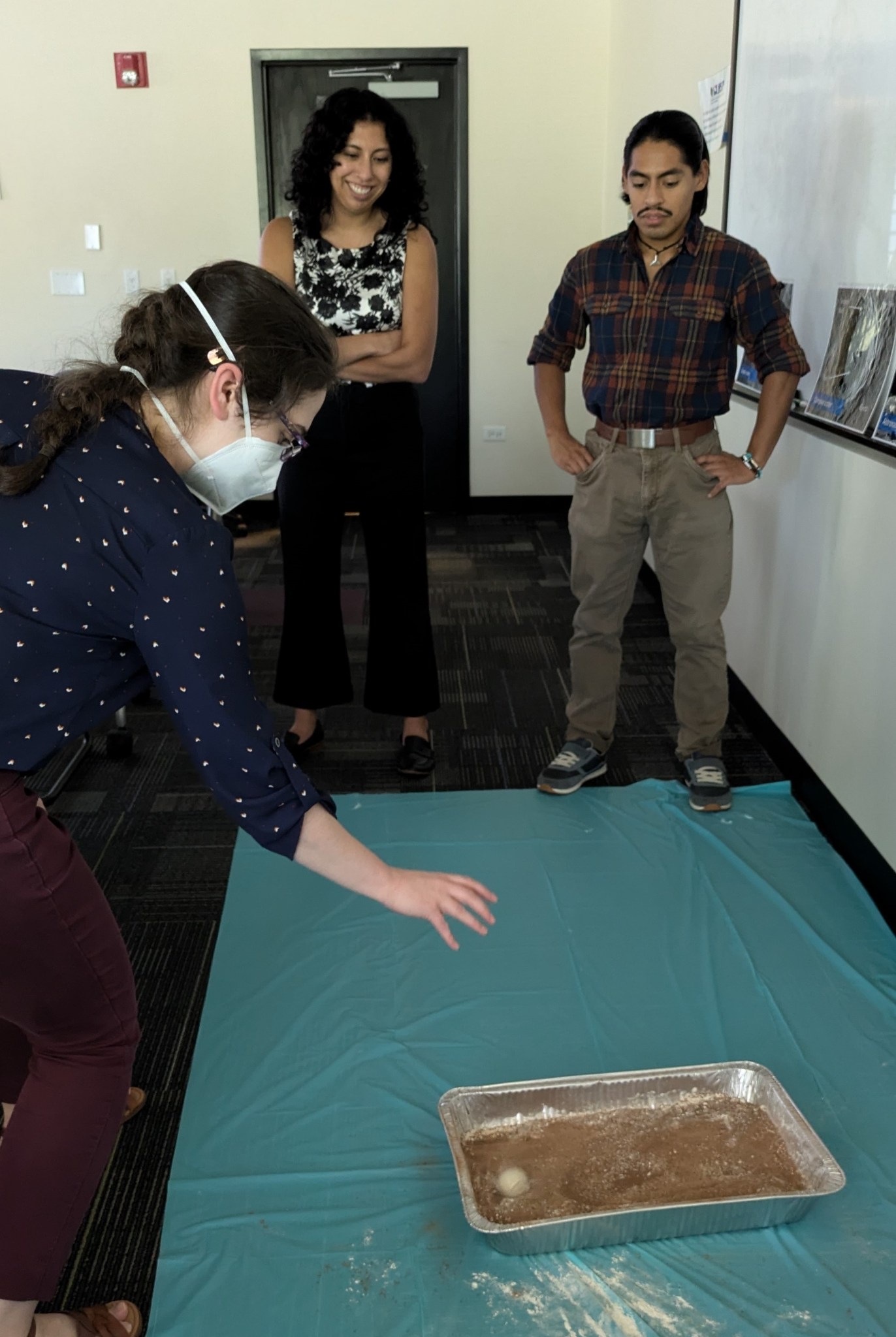 A standing adult who appears to have dropped a ball leans over a large pan on the floor, which holds different colored materials and the ball. Two smiling adults in the background watch.